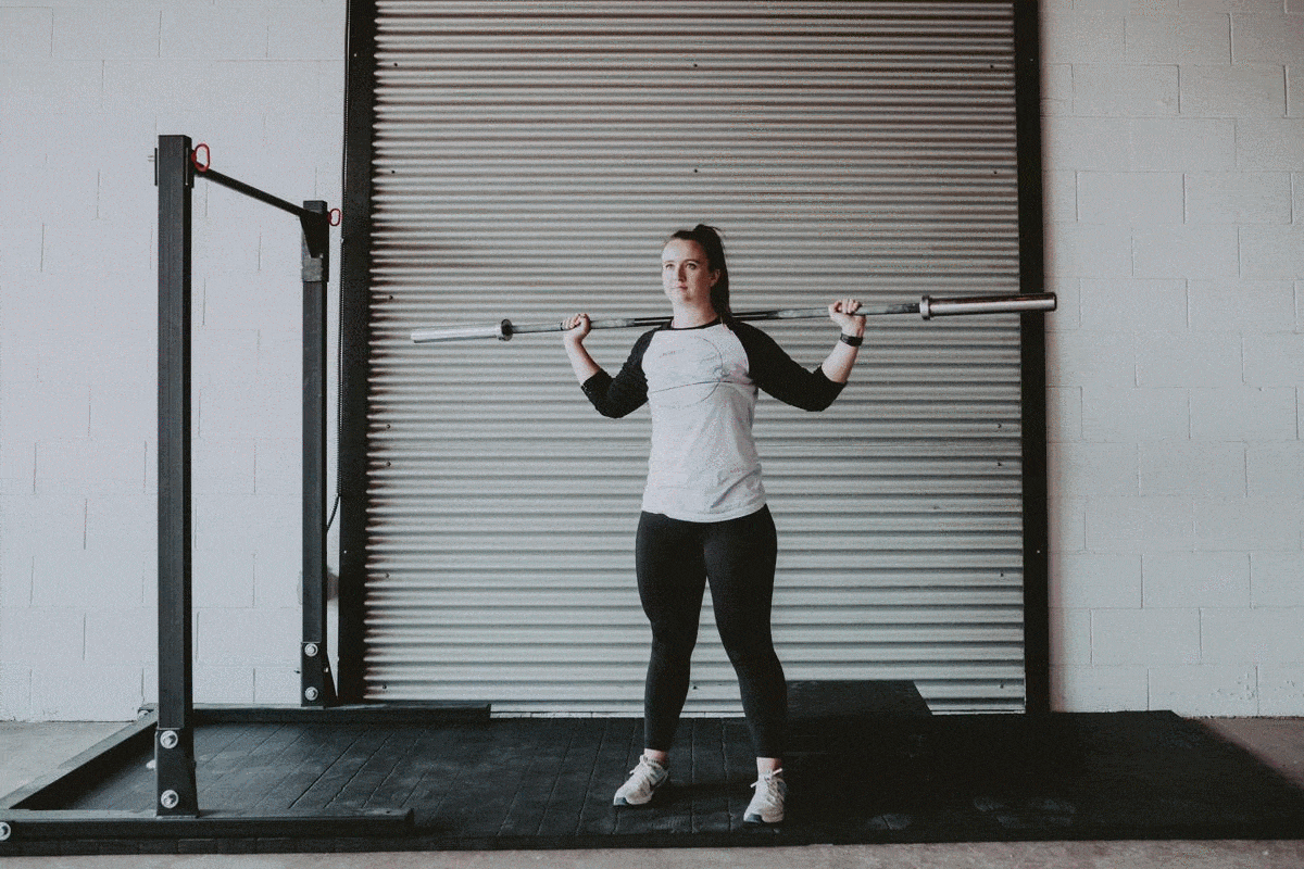 Girl performs a squat with barbell. The girl is wearing a black and white baseball T with Victoria BC CrossFit Gym CrossFit LoLo Logo on the front